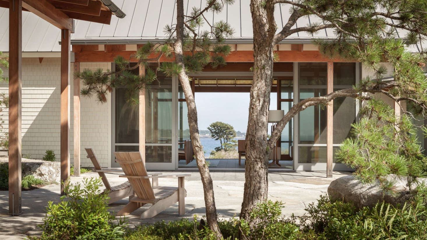 Bordered by full-height sliding glass doors, the living room links a courtyard in front of the house to a patio on the opposing side. The landscape architect planted pitch pine here, centered on the axis of a single large pitch pine hovering over the water