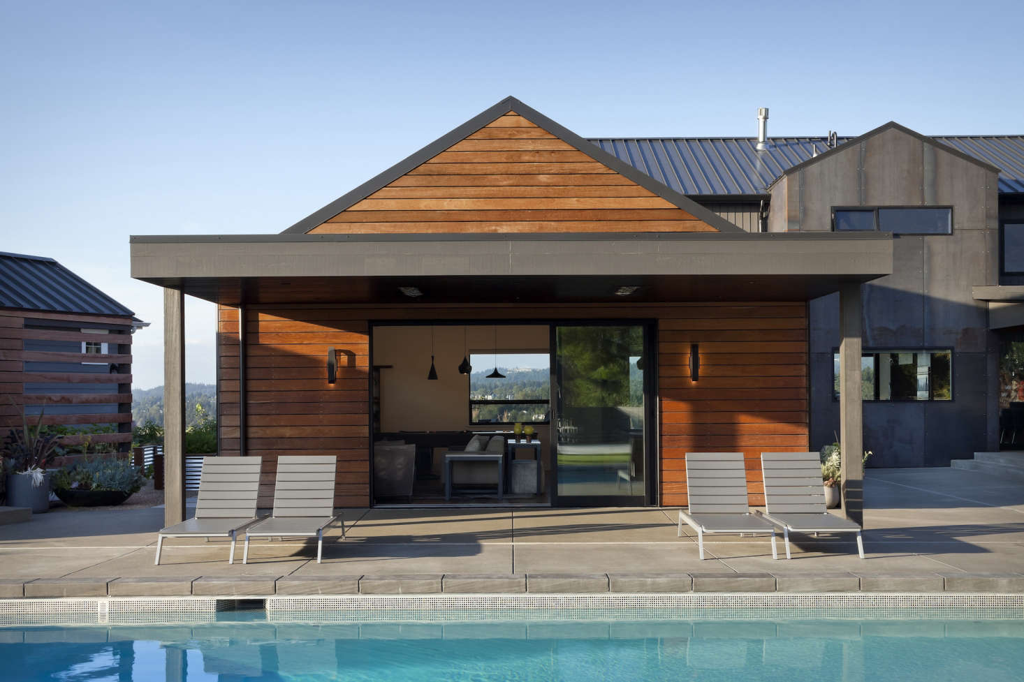 A media room is attached to the house but functions as an indoor-outdoor space during parties on the patio. Sliding glass doors open onto the pool deck, and a flat overhang creates shade for lounge chairs. The overhang and trim is painted in Iron Ore from Sherwin-Williams.