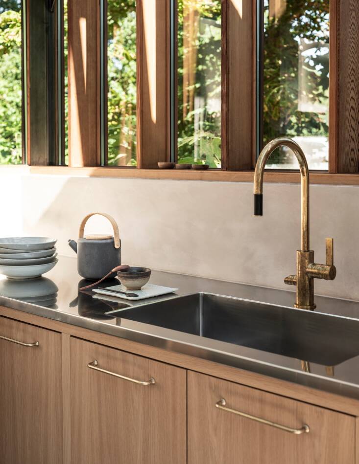 Light oak cabinets, antique brass hardware by Corston, and a brass faucet by Tapwell are warm counterpoints to the stainless steel countertops and appliances.